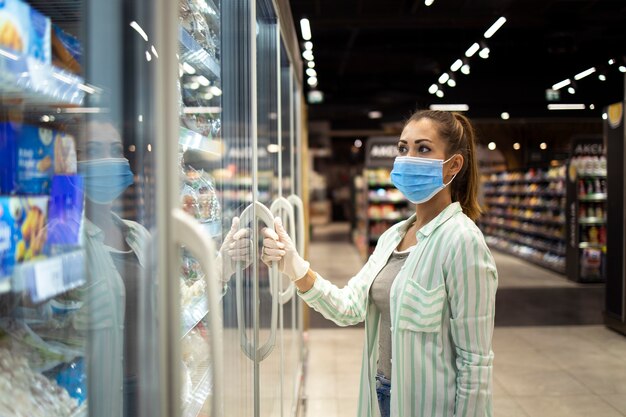 Mujer con máscara protectora y guantes abriendo congelador en supermercado durante la pandemia COVID-19 o el virus corona