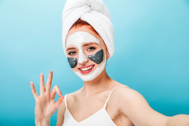 Mujer con mascarilla mostrando signo bien y riendo. Modelo femenino tomando selfie mientras hace un tratamiento de spa.