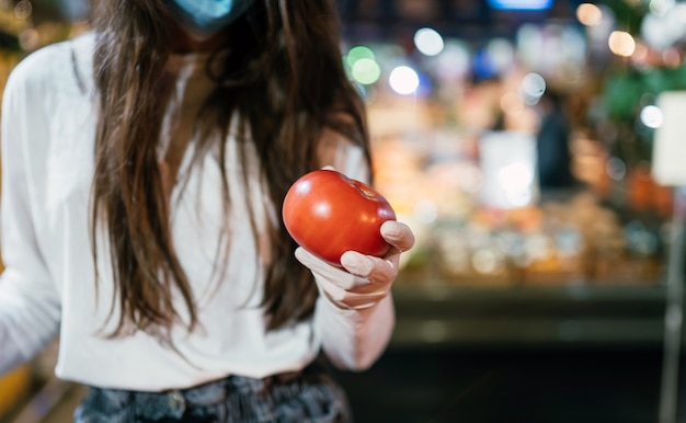 Mujer con mascarilla quirúrgica y guantes está comprando en el supermercado después de la pandemia de coronavirus. la chica de la mascarilla va a comprar tomates.