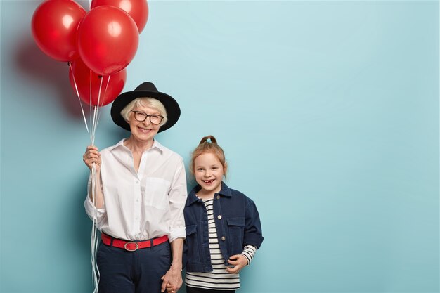 La mujer mayor alegre y la nieta se dan la mano, tienen una actitud positiva, expresiones faciales alegres, viste un atuendo elegante, vamos al evento festivo dedicado al Día del Niño. Dos generaciones