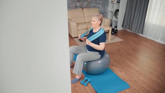 Mujer mayor alegre que ejercita en la bola del balance. Entrenamiento de personas mayores en el hogar, deporte, estilo de vida saludable, ejercicios de fitness para ancianos en el apartamento, actividad y atención médica