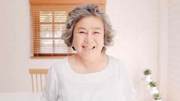 Mujer mayor asiática que siente la sonrisa feliz y que mira a la cámara mientras que relájese en el sofá en sala de estar en casa. estilo de vida de las mujeres mayores en el concepto de hogar.