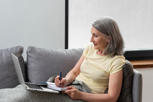Mujer mayor en casa en el sofá usando la computadora portátil y tomando notas