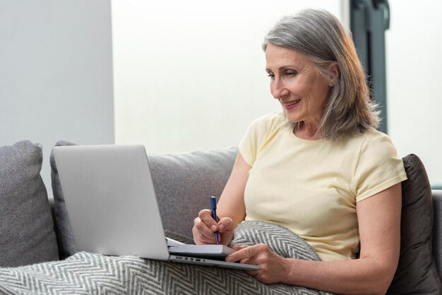 Mujer mayor en casa en el sofá usando la computadora portátil y tomando notas