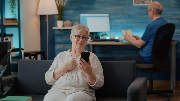 Mujer mayor chateando en una videollamada remota en la sala de estar. Adulto mayor que usa teléfono móvil para conversar en videoconferencia en línea para telecomunicaciones. Reunión de Internet en el teléfono inteligente