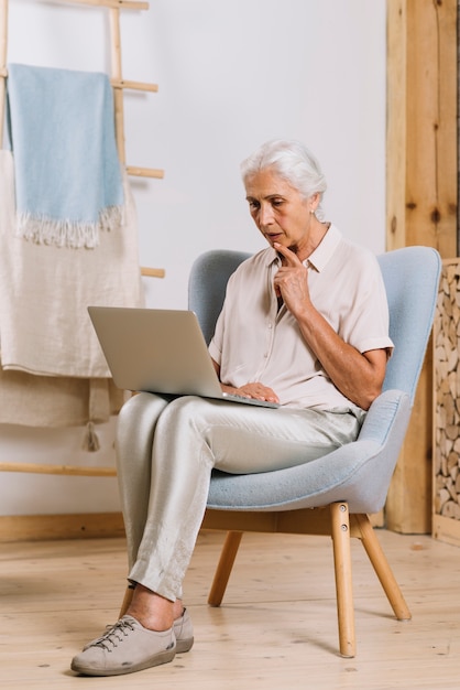 Mujer mayor contemplada que se sienta en la butaca que mira la computadora portátil