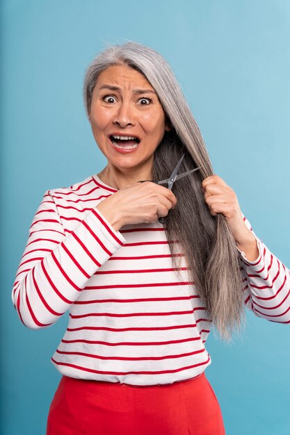 Mujer mayor cortándose el pelo y asustada contra un fondo azul