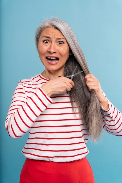 Mujer mayor cortándose el pelo y asustada contra un fondo azul