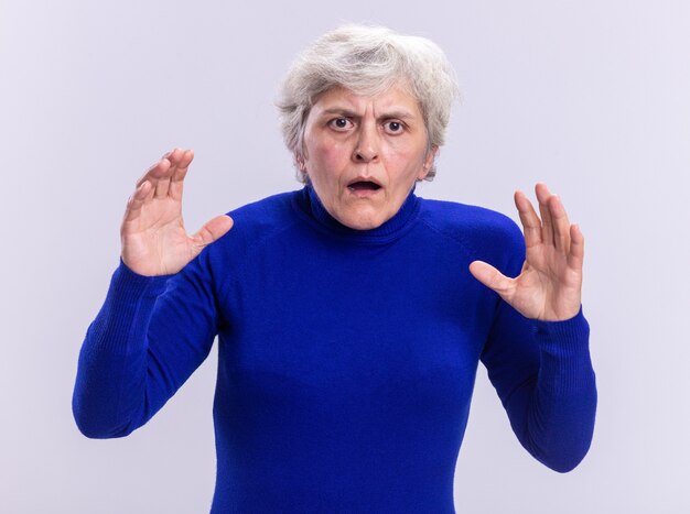 Mujer mayor en cuello alto azul mirando a la cámara preocupada y confundida con los brazos levantados de pie sobre fondo blanco.