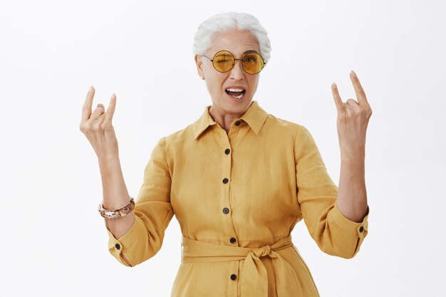 Mujer mayor despreocupada con estilo en gafas de sol mostrando gesto de rock-n-roll y sonriendo, divirtiéndose