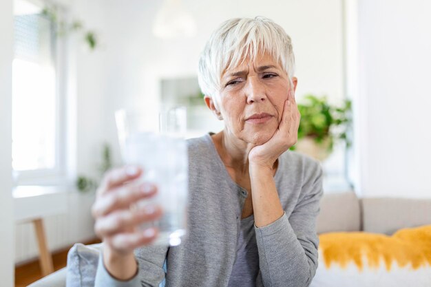 Mujer mayor con dientes sensibles y mano sosteniendo un vaso de agua fría con hielo Concepto de salud Mujer madura bebiendo un vaso de bebida fría lleno de cubitos de hielo y siente dolor de muelas