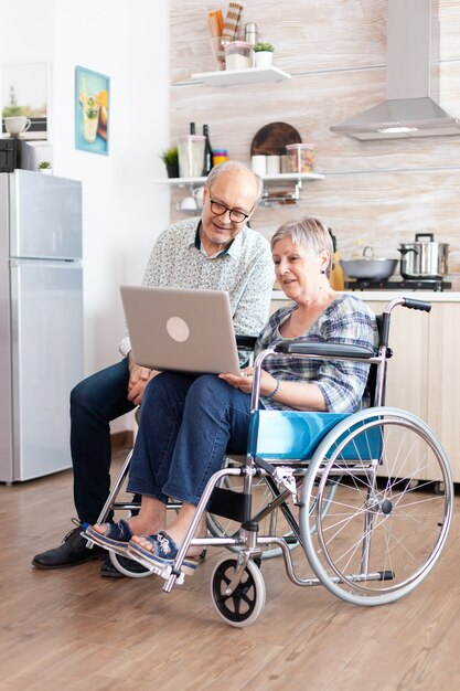 Mujer mayor discapacitada en silla de ruedas y su esposo buscando en la computadora portátil, navegando en las redes sociales sentado en la cocina por la mañana. persona mayor discapacitada paralizada que tiene una conferencia en línea.