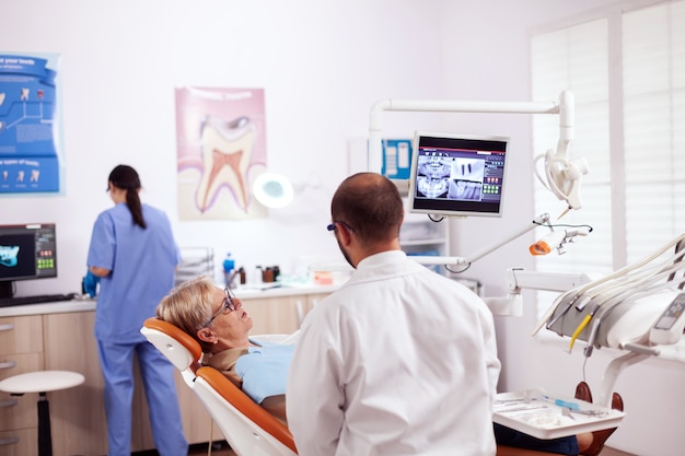 Mujer mayor discutiendo con el dentista en el gabinete dental sobre la cuestión de los dientes sentado en una silla. Tomador del cuidado de los dientes médicos discutiendo con la mujer mayor acerca de la higiene bucal.