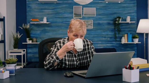 Mujer mayor disfrutando de una taza de café mientras trabaja en la computadora portátil en la sala de estar mientras su esposo está sentado en el sofá y leyendo un libro en segundo plano. Señora mayor madura viendo formación empresarial en línea