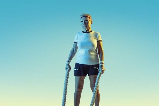 Mujer mayor en entrenamiento de ropa deportiva con cuerdas sobre fondo degradado, luz de neón.