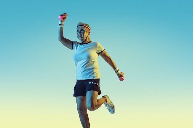 Mujer mayor en entrenamiento de ropa deportiva con pesas sobre fondo degradado, luz de neón. Modelo femenino en gran forma se mantiene activo. Concepto de deporte, actividad, movimiento, bienestar, confianza. Copyspace.