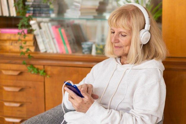 Mujer mayor escuchando música en casa con smartphone y auriculares