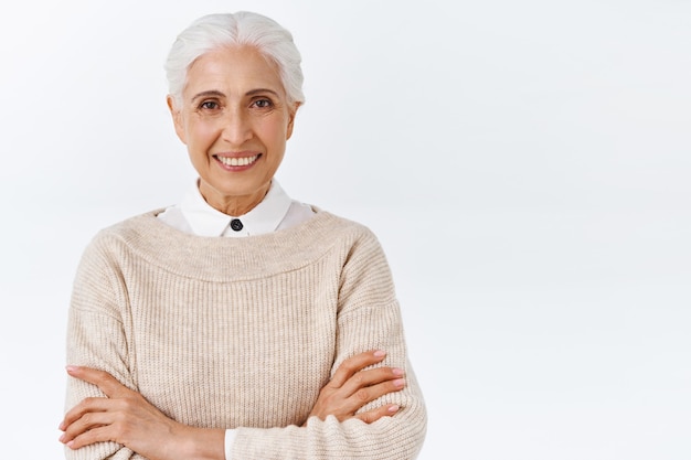 Foto gratuita mujer mayor feliz y complacida segura de sí misma con corte de pelo peinado gris, brazos cruzados sobre el pecho como profesional, de pie, determinada pared blanca