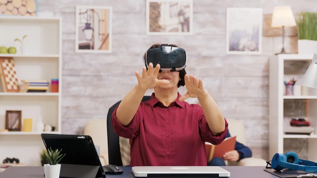 Mujer mayor jubilada asombrada mientras usa gafas de realidad virtual en la sala de estar. Anciano sentado en el sofá mientras lee un libro.