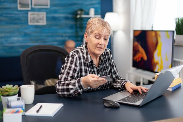 Mujer mayor leyendo el código CVV de la tarjeta de crédito sentado frente a la computadora portátil