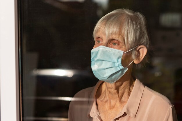 Mujer mayor con máscara médica mirando por la ventana