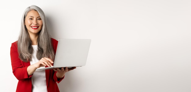 Foto gratuita mujer mayor de negocios que trabaja en un portátil con ropa de oficina y sonriendo de pie sobre el fondo blanco