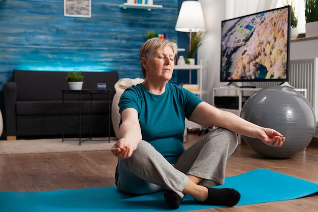 Mujer mayor pacífica con los ojos cerrados sentado en la estera de yoga meditando durante el entrenamiento de bienestar. Cómodo pensionista practicando la posición de loto ejercitando la concentración corporal en el salón