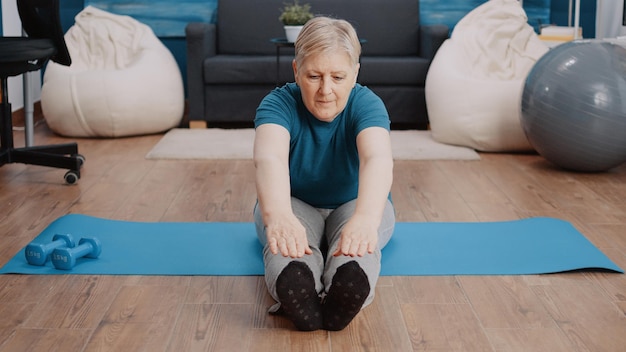 Mujer mayor que se extiende en posición de aeróbic en la estera de yoga. Persona mayor que hace ejercicio físico y entrena para el bienestar y la atención médica. Pensionista haciendo ejercicio de gimnasia en casa.