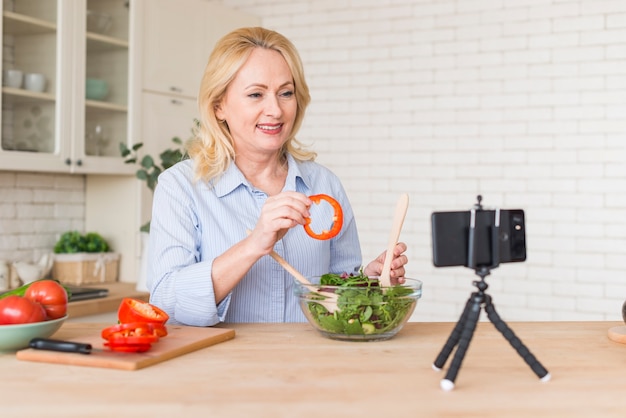 Mujer mayor que hace la llamada video en el teléfono móvil que muestra la rebanada del paprika mientras que prepara la ensalada