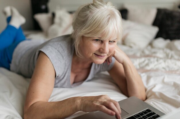 Mujer mayor que hojea la computadora portátil en cama