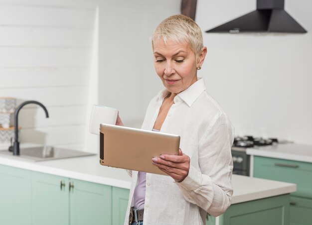 Mujer mayor que mira en su tableta en la cocina