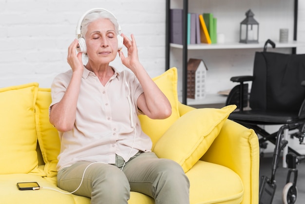 Mujer mayor que se sienta en la música que escucha del sofá amarillo en los auriculares