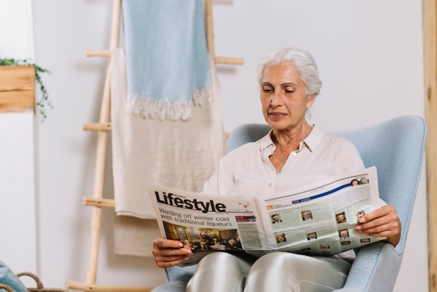 Mujer mayor que se sienta en periódico de la lectura de la silla