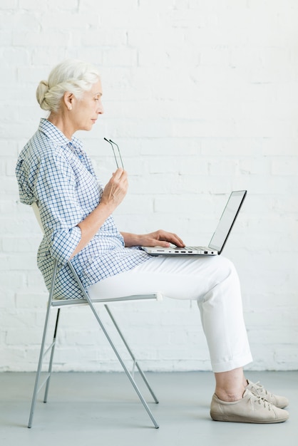 Mujer mayor que se sienta en silla usando la computadora portátil