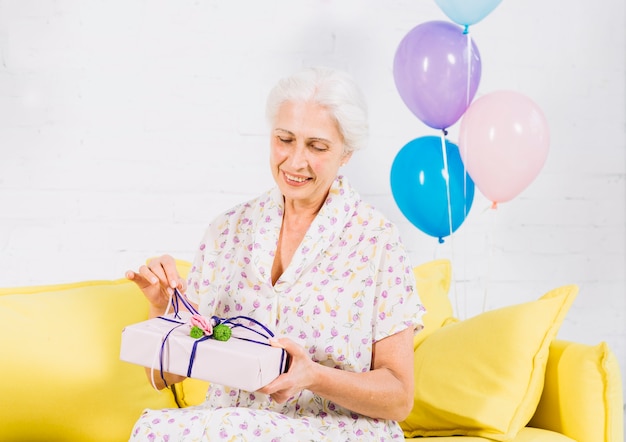 Mujer mayor que se sienta en el sofá que desempaqueta el regalo de cumpleaños
