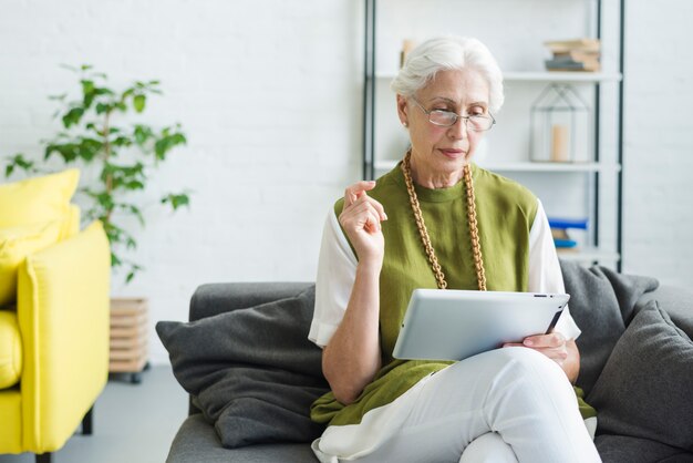 Mujer mayor que se sienta en el sofá que mira la tableta digital