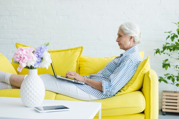 Mujer mayor que se sienta en el sofá usando la computadora portátil