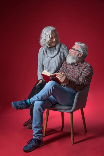 Mujer mayor que se sienta con su marido que se sienta en la silla que sostiene el libro disponible contra fondo rojo