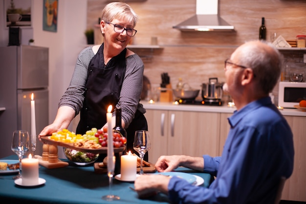 Mujer mayor que sostiene la placa de madera con y mirando al marido durante la cena festiva. Pareja de ancianos hablando, sentados a la mesa en la cocina, disfrutando de la comida, celebrando su aniversario.