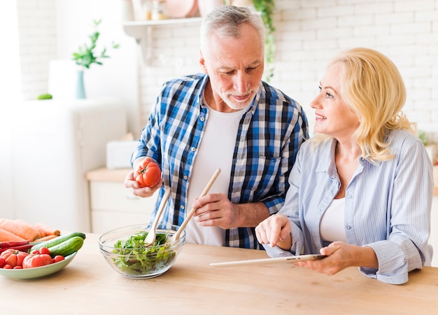 Mujer mayor que sostiene la tableta digital en la mano que muestra receta a su marido que prepara la ensalada en la cocina