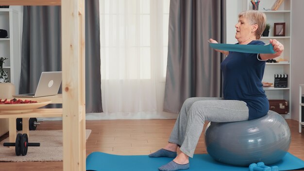 Mujer mayor que trabaja con bola de equilibrio y banda de resistencia. Entrenamiento de personas mayores en el hogar, deporte, estilo de vida saludable, ejercicios de fitness para ancianos en el apartamento, actividad y atención médica