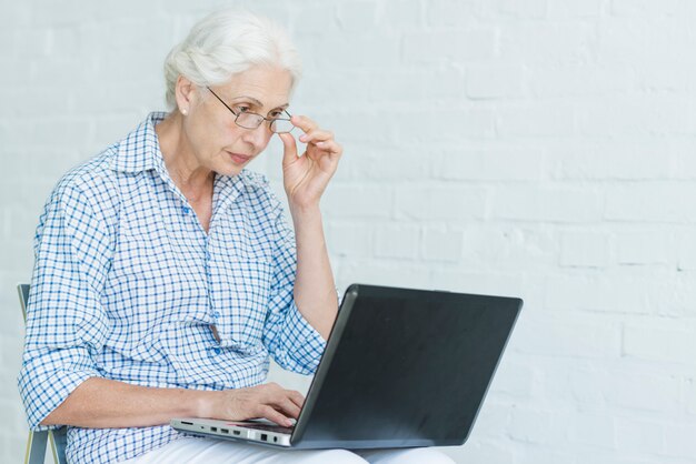 Mujer mayor que usa la computadora portátil contra la pared blanca