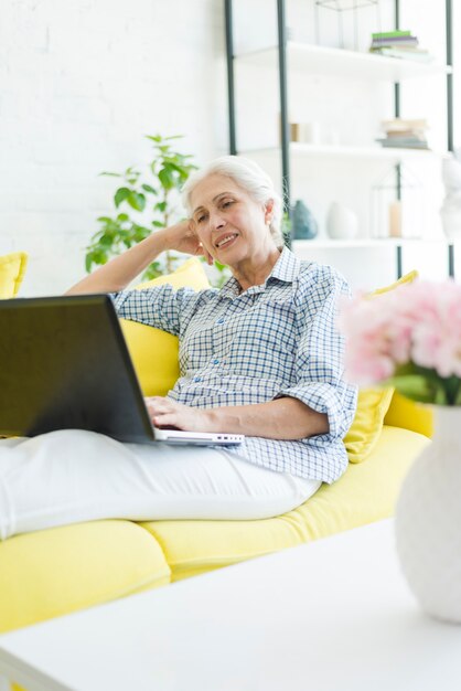 Mujer mayor relajada que se sienta en el sofá que mira la computadora portátil