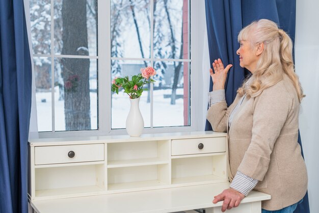 Mujer mayor rubia que mira a través de la ventana en casa