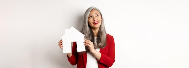Mujer mayor soñadora pensando en comprar una propiedad que muestra el recorte de la casa de papel y mira la parte superior izquierda