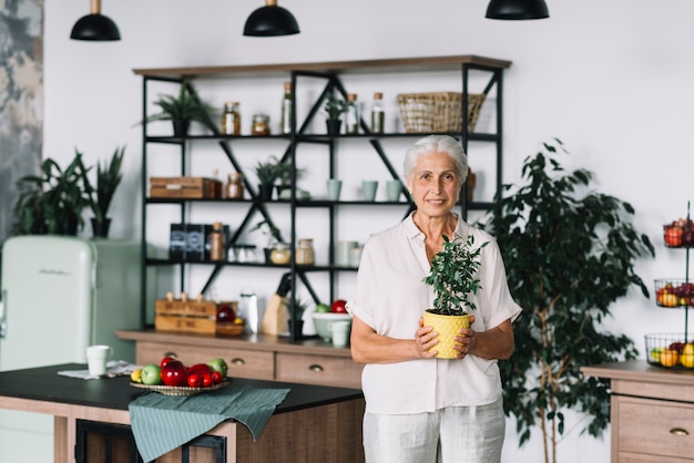 Mujer mayor sonriente que se coloca en la cocina que sostiene la planta de tiesto