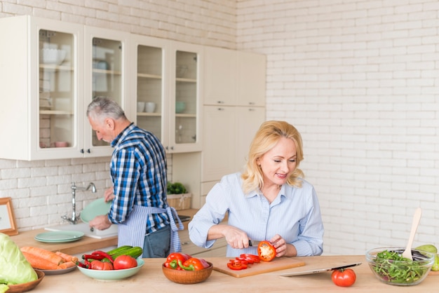 Foto gratuita mujer mayor sonriente que corta el pimiento rojo con el cuchillo que mira la tableta digital y su marido que lavan platos en el fregadero de cocina