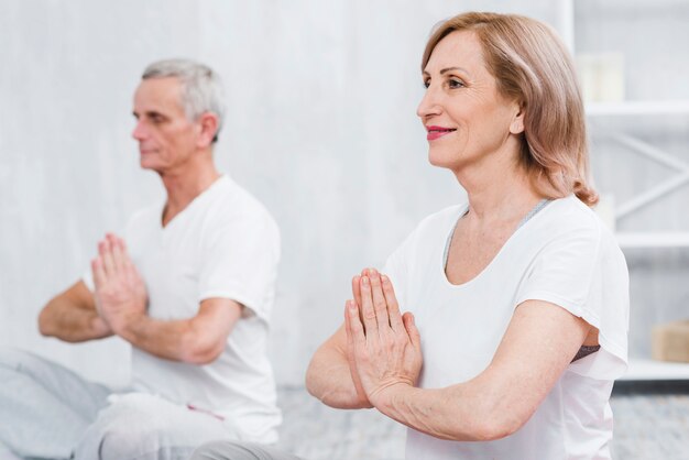 Mujer mayor sonriente que hace yoga con gesto de mano de rogación en hogar