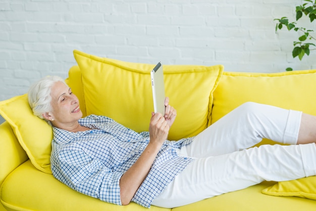 Mujer mayor sonriente que miente en el sofá amarillo que mira la tableta digital