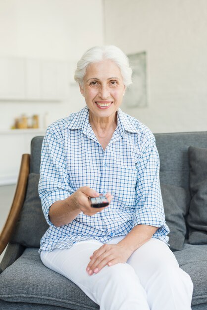 Mujer mayor sonriente que se sienta en el sofá usando teledirigido